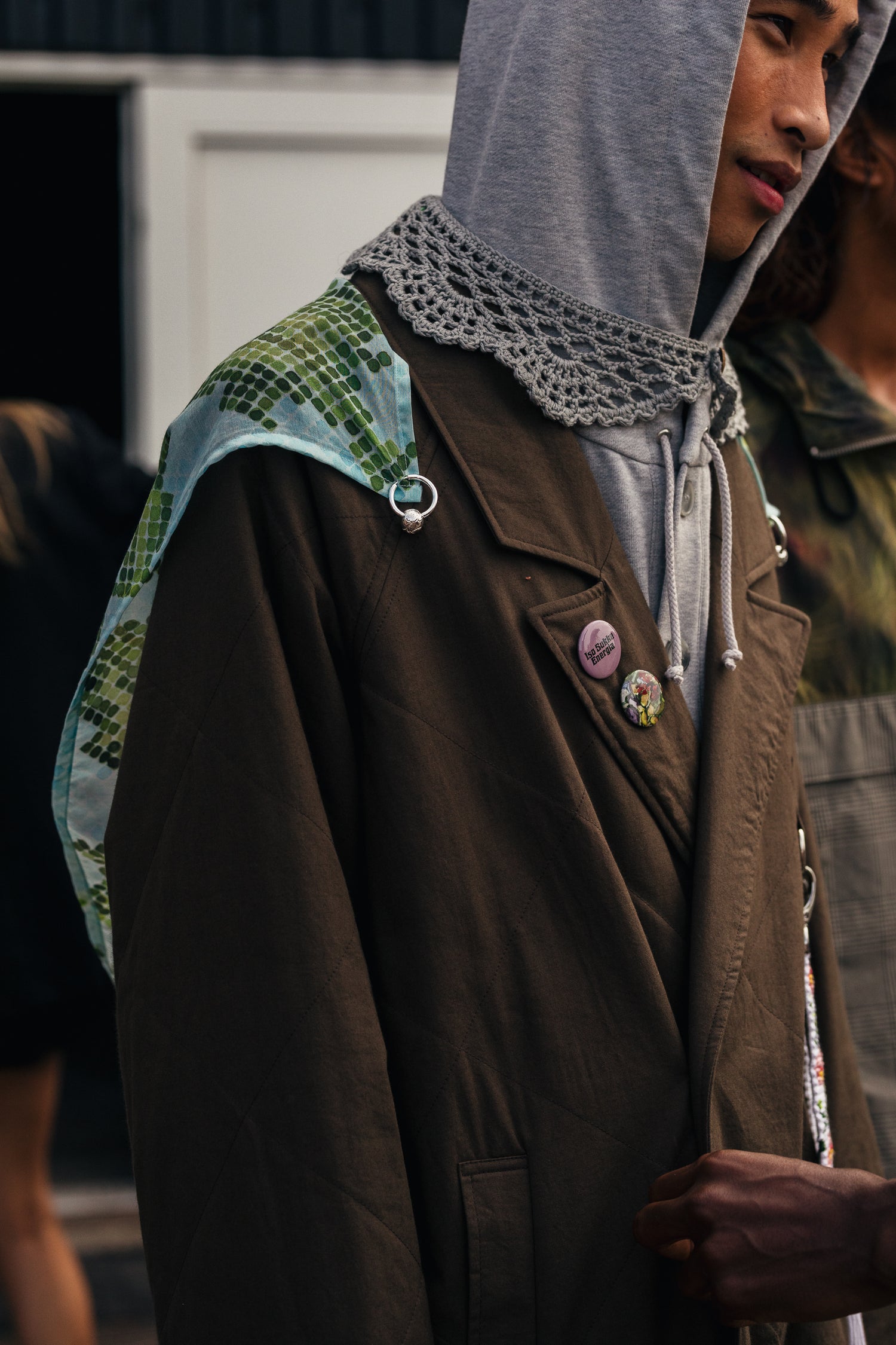 Mallin ruskeaan takkiin on kiinnitetty RAKAS Rolf Ekroth x Kalevala -koruilla ruusukuvioinen huivi. Lisäksi hänellä on harmaa huppari ja virkattu kaulus.